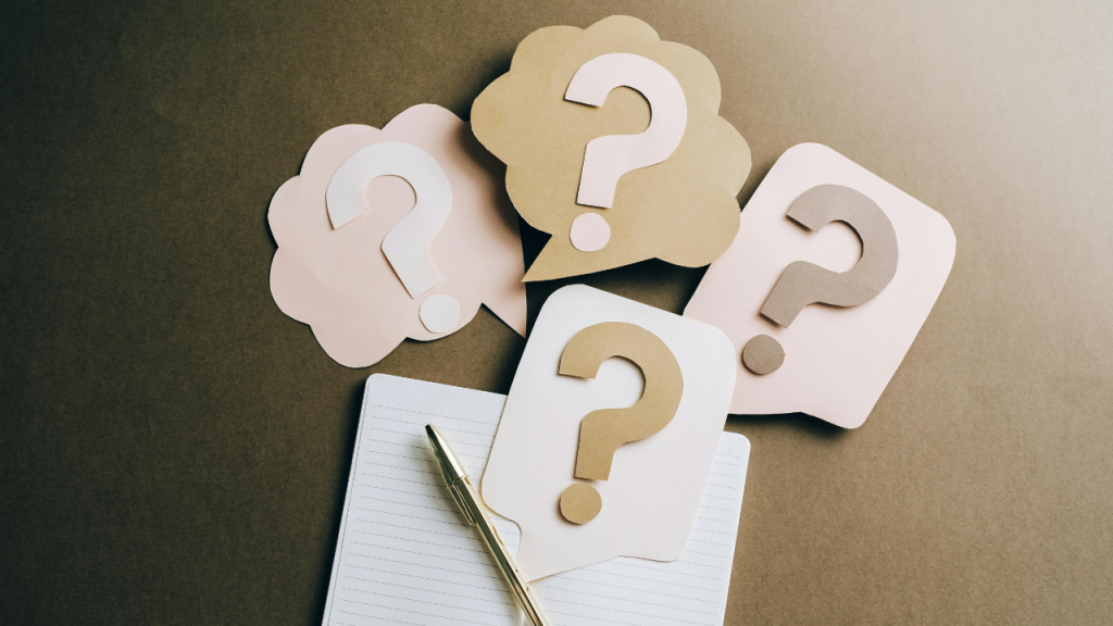 Multiple pastel-colored question marks on paper cutouts above an open notebook and pen, symbolizing FAQs many face when starting an ADHD skincare routine